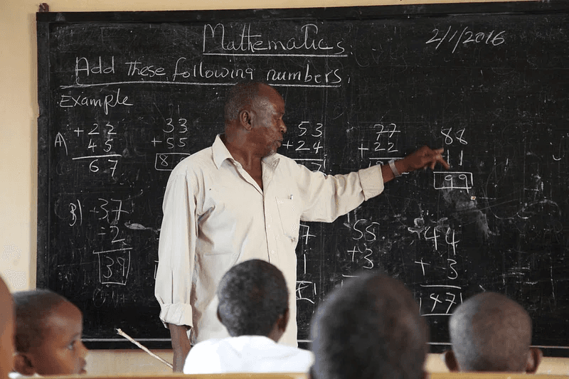 Mathematics educator pointing to sums on the chalkboard while learners look on.